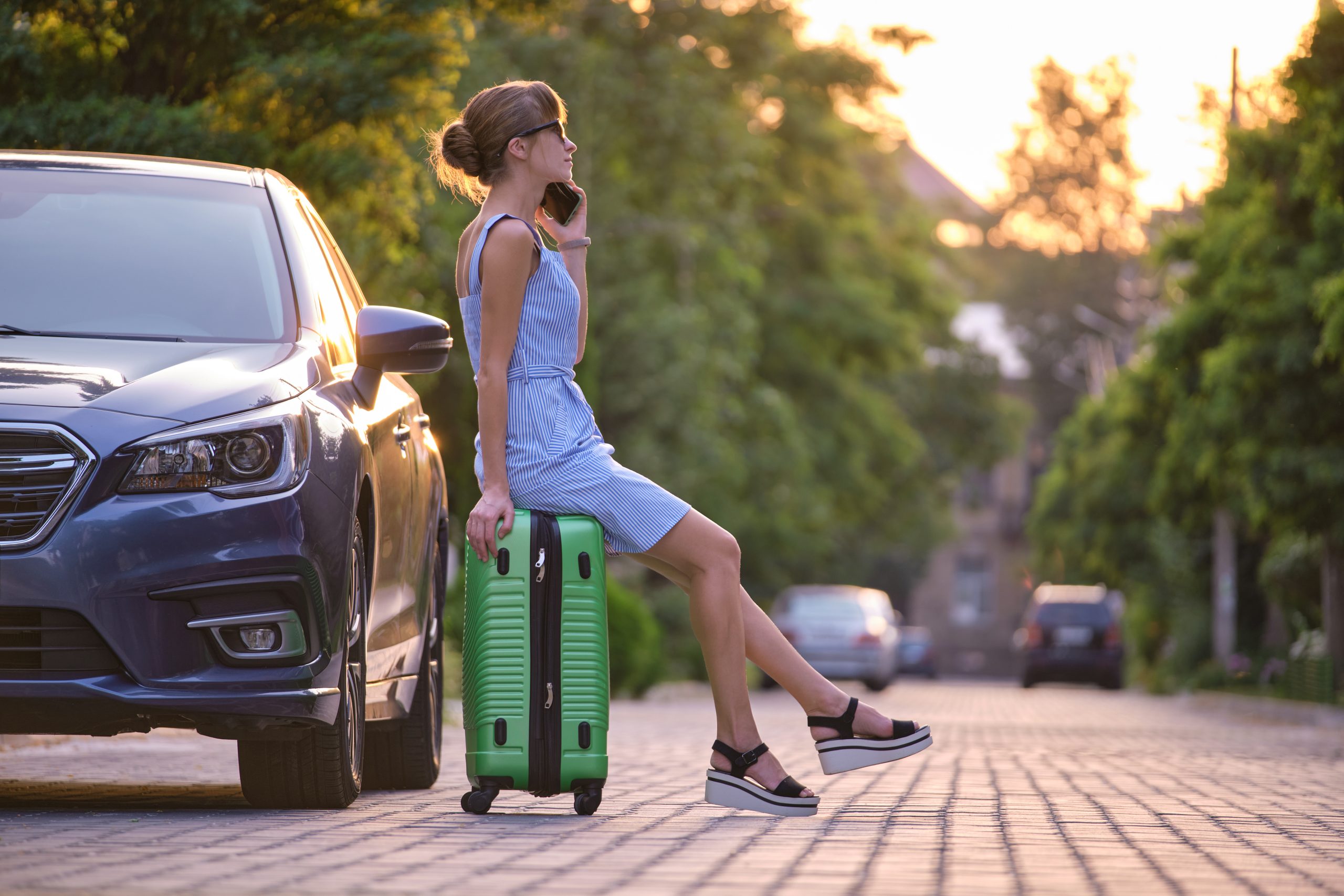 Young female driver sitting on suitcase near her car speaking on Hassle-Free Travel